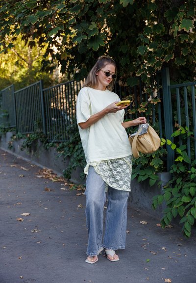 Camiseta oversized verde claro, falda con detalles de encaje sobre jeans azul claro, bolso cruzado beige y sandalias planas. Gafas de sol oscuras completan el conjunto.