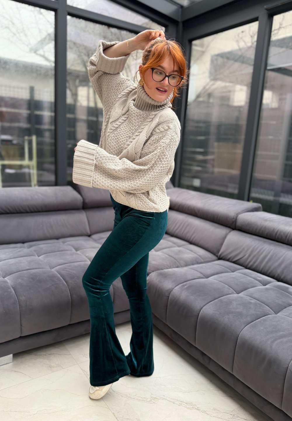 Femme aux cheveux roux portant des lunettes, un pull beige et un pantalon vert foncé, posant dans un salon moderne aux murs en verre avec des canapés gris.