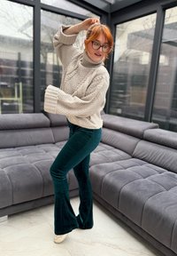 Femme aux cheveux roux portant des lunettes, un pull beige et un pantalon vert foncé, posant dans un salon moderne aux murs en verre avec des canapés gris.