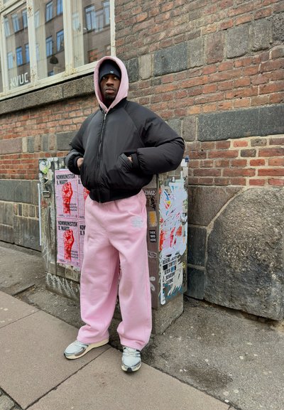 Hombre con chaqueta acolchada negra, sudadera con capucha rosa y pantalones deportivos, de pie con las manos en los bolsillos cerca de una pared de ladrillos cubierta de carteles y pegatinas.