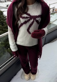 Suéter de punto acogedor en color marfil con diseño de lazo granate, combinado con guantes y pantalones de punto con cableado en color granate. Zapatos de color crema completan el conjunto.