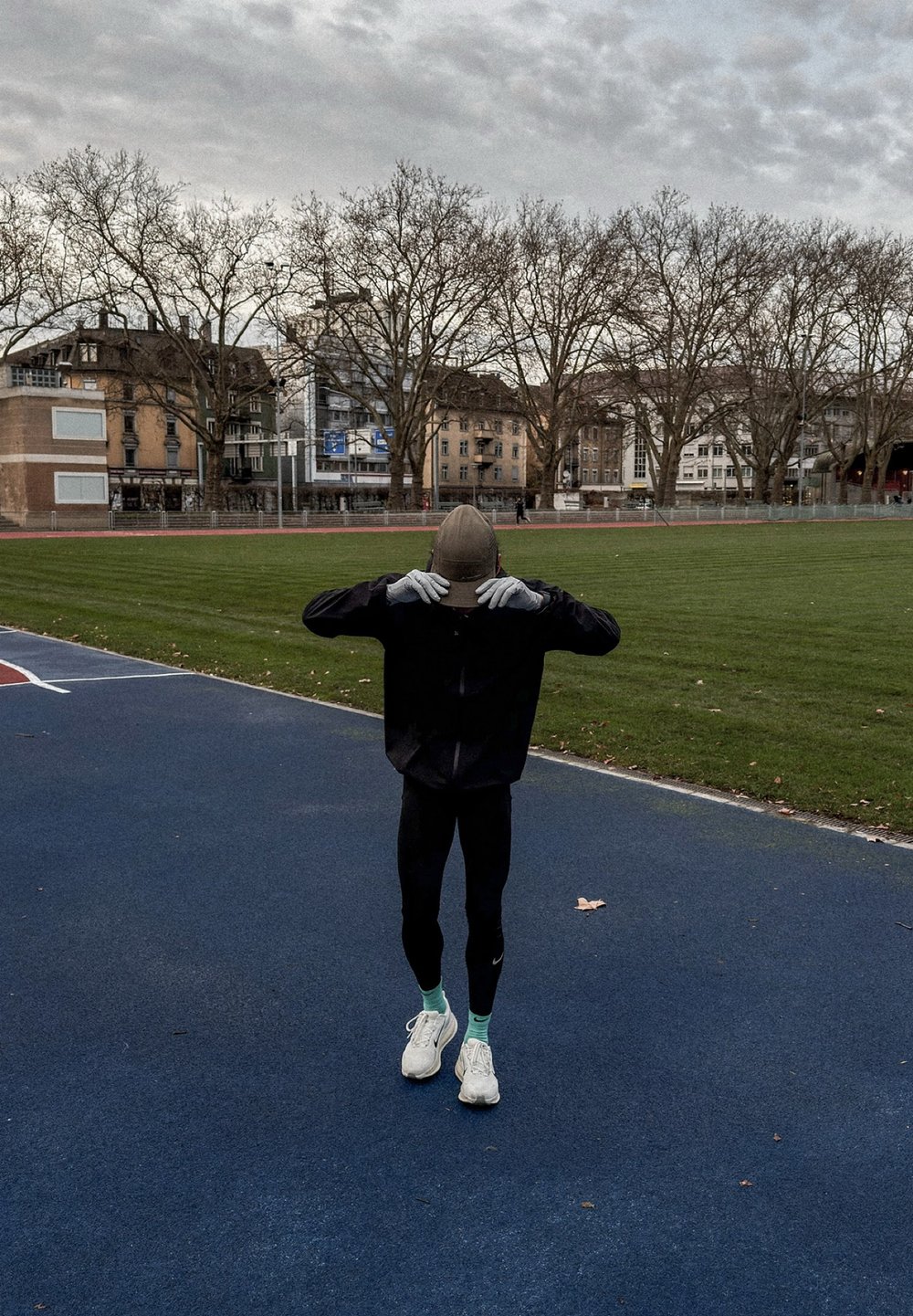 Egy sportoló fekete dzsekit, szoros fekete leggingset, fehér cipőt és szürke kesztyűt visel, és bézs színű sapkát igazít egy kék futópályán, a fű közelében.