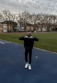 Egy sportoló fekete dzsekit, szoros fekete leggingset, fehér cipőt és szürke kesztyűt visel, és bézs színű sapkát igazít egy kék futópályán, a fű közelében.