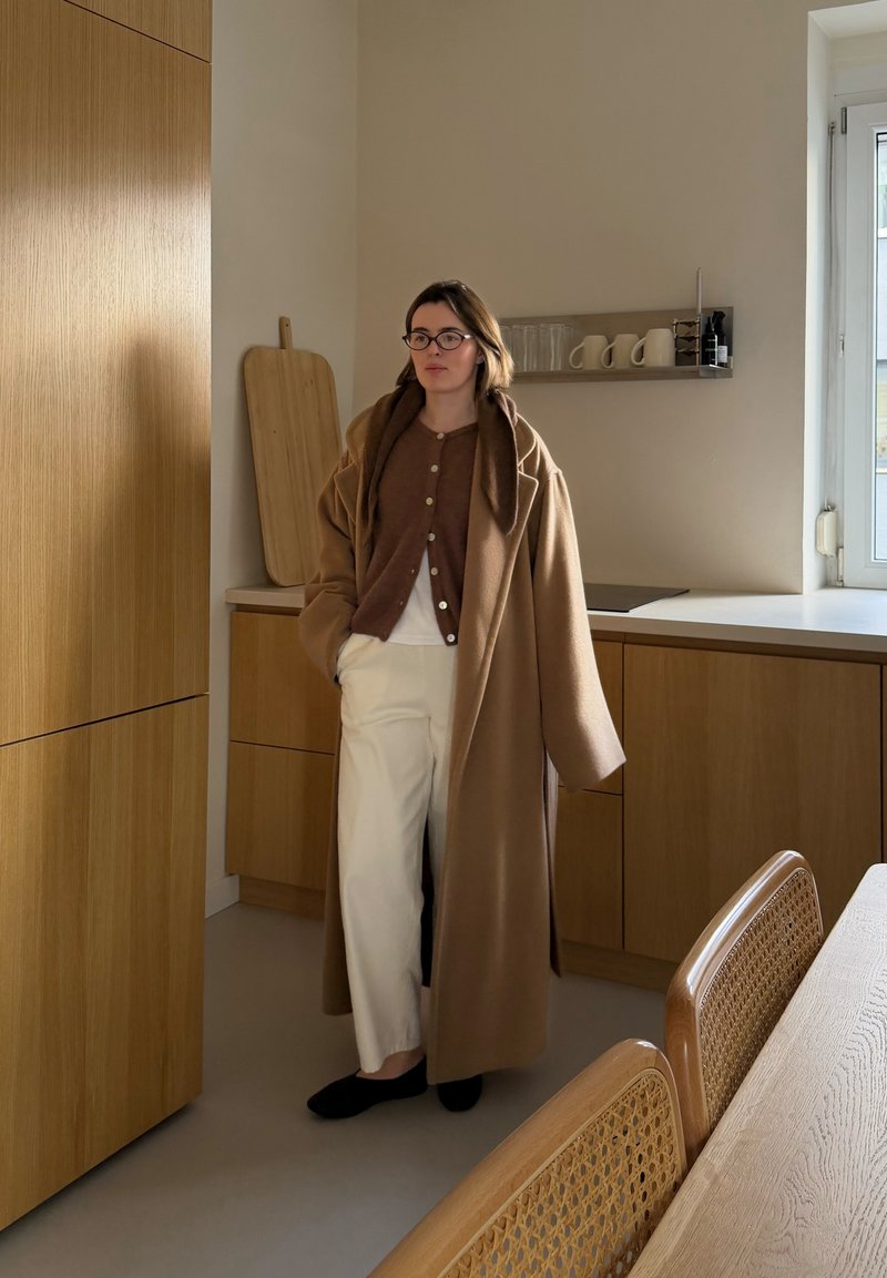 Femme portant un long manteau marron, un cardigan marron, un pantalon blanc et des lunettes, debout dans une cuisine moderne avec des armoires en bois et une fenêtre.