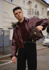 Muž oblečený v lesklej tmavočervenej koženej bunde a čiernych nohaviciach drží trojuholníkovú tmavo hnedú kabelku na ulici pred budovou.