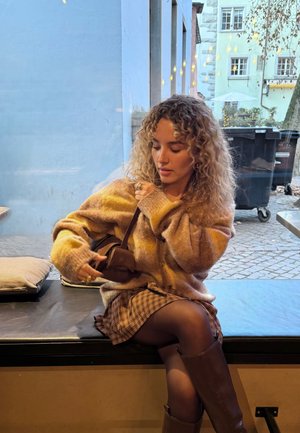 Mujer con cabello rizado ajustando un bolso pequeño marrón, vestida con un suéter beige, falda de cuadros y botas marrones, sentada junto a una ventana en una cafetería.