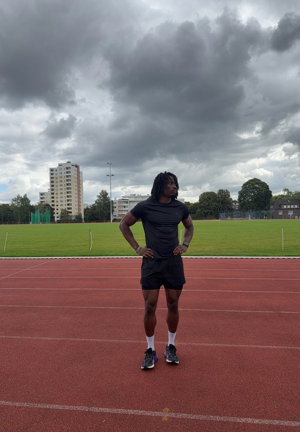 Athlète en tenue de sport noire debout sur une piste rouge avec les mains sur les hanches, champ vert et ciel nuageux en arrière-plan.
