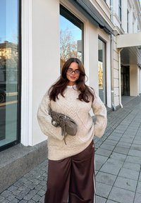 Lysebeige strikket sweater med kabelmønster, kombineret med mørkebrune bukser med vide ben. Holder en grå pungtaske. Urban baggrund med vinduer.