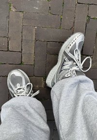 Grå New Balance-sneakers med ett nätmönster, utrustade med en vadderad sula, snören och en distinkt "N"-logotyp, tillsammans med grå byxor på asfalt.