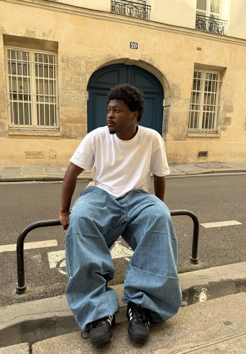 Jean en denim bleu clair large, t-shirt blanc, baskets noires. La scène montre une rue avec un bâtiment beige et une porte bleu foncé.