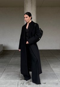 Manteau en laine noire à coupe ajustée, manches longues et fermeture à un bouton, associé à un chemisier noir et un pantalon à jambes larges.