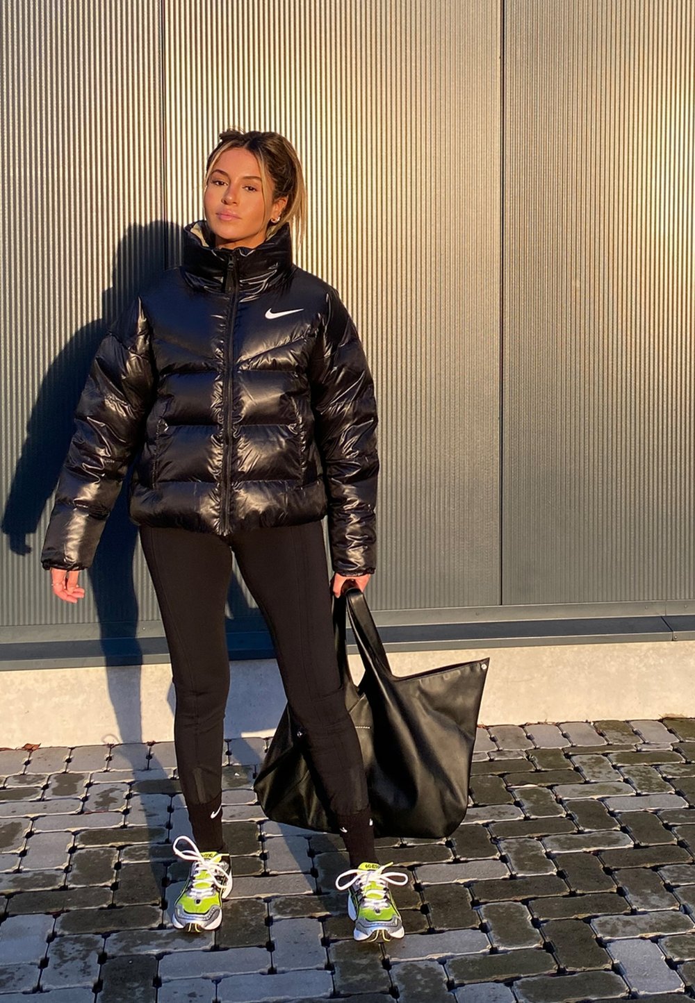 Mulher jovem com uma jaqueta puffer preta, leggings pretas e ténis neon est�á de pé numa pavimentação de azulejos, segurando uma grande carteira de mão preta.