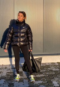 Mulher jovem com uma jaqueta puffer preta, leggings pretas e ténis neon está de pé numa pavimentação de azulejos, segurando uma grande carteira de mão preta.