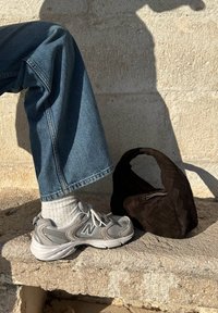 Harmaat New Balance -lenkkarit, valkoiset sukat, siniset väljät farkkutalot ja musta neulottu käsilaukku kaarevalla kahvalla kivipinnalla.