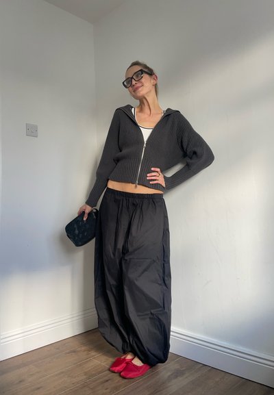 Mujer posando en interiores con pantalones negros holgados, suéter corto con cierre, top blanco, gafas negras, zapatos planos rojos y sosteniendo una pequeña bolsa negra.