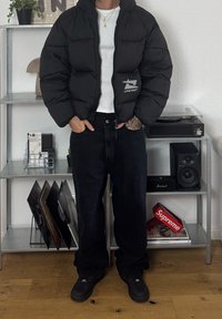 Inimene, kandes musta puffjakki, millel on "Live Fast" logo, valge särk, mustad teksad ja mustad kingad, seisab siseruumides metallshelfi lähedal, kus on vinüülplaadid.