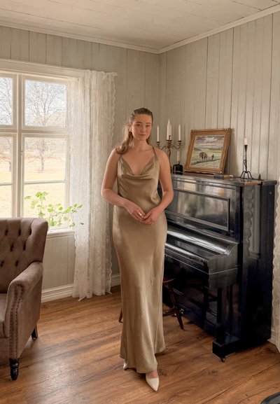 Mujer con un vestido dorado de seda junto a un piano vertical negro en una habitación soleada con suelos de madera, paredes beige, cortinas de encaje y un sillón marrón.