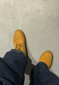 Cizme de lucru galbene cu șireturi, confecționate din piele, cu o textură ușor uzată. Asortate cu blugi denim albastru închis pe o suprafață gri deschis.