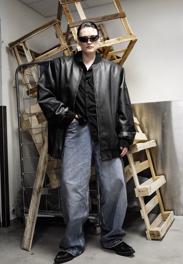 Zwarte lederen oversized bomberjack, zwart shirt, losse blauwe spijkerbroek en zwarte schoenen, met een zonnebril en houten pallets op de achtergrond.