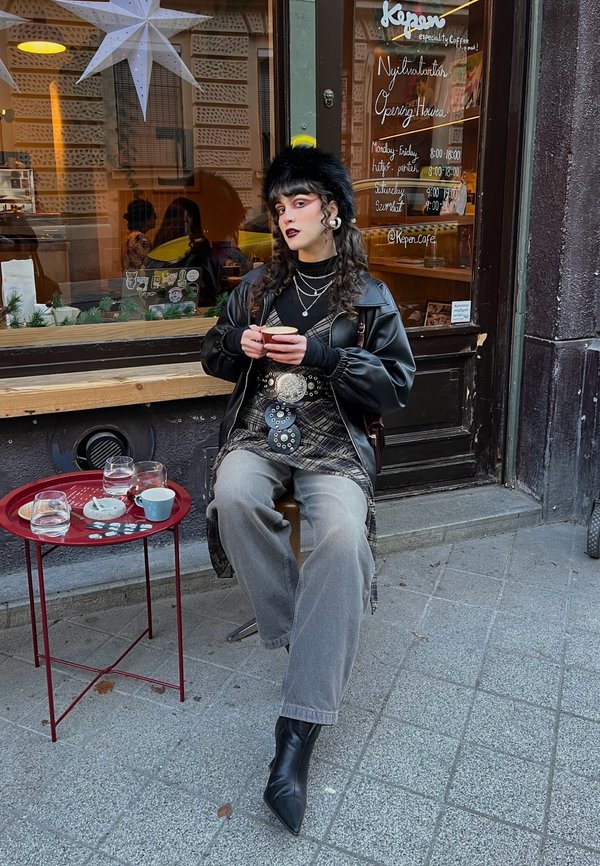 Mlajša oseba s kodrastimi lasmi in temnim šminko sedi zunaj kavarne, drži skodelico, oblečena v črno usnjeno jakno, škornje in krzneno kapo.