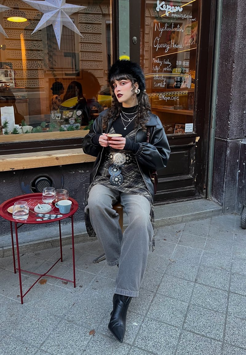 Jeune personne aux cheveux bouclés et rouge à lèvres foncé assise à l'extérieur d'un café, tenant une tasse, portant une veste en cuir noire, des bottes et un chapeau en fourrure.