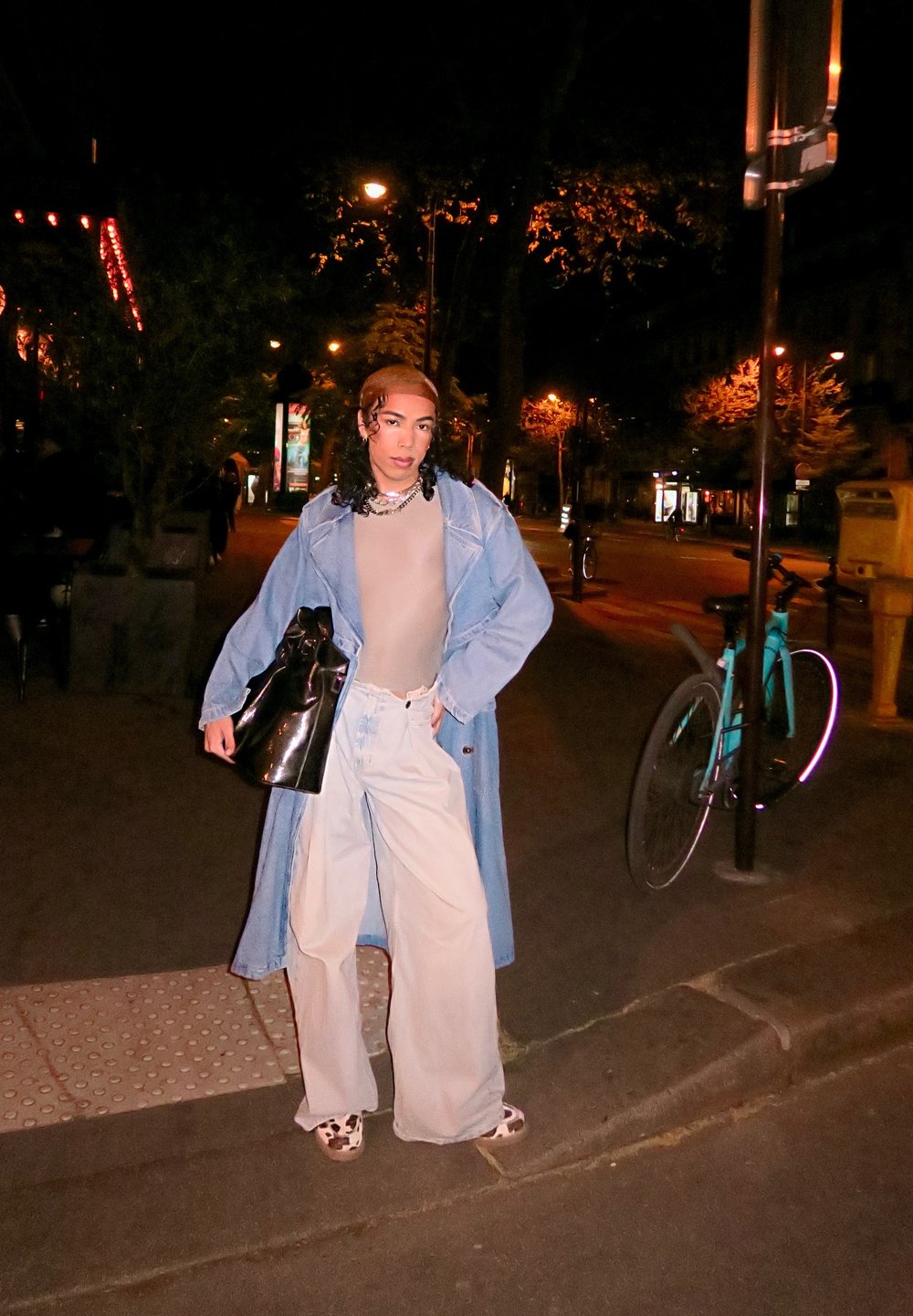 Persoon in een denim trenchcoat, beige top, brede witte broek en schoenen met koeienprint staat 's nachts op een stadsstoep met een zwarte tas in de hand.