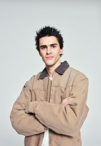 Young man with spiky dark hair wearing a beige jacket, standing with folded arms against a plain light grey background.