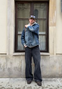Jeansjacka över grå hoodie, svarta vida byxor, bruna skor. Tillbehör inkluderar en keps och solglasögon. Stående mot en byggnad.