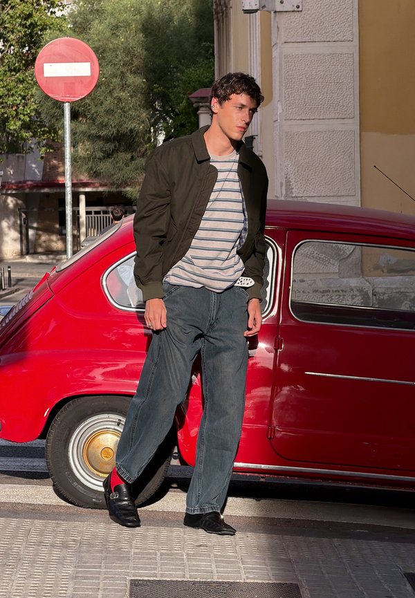 Joven con camisa de rayas y chaqueta oscura caminando junto a un coche antiguo rojo cerca de una señal de tráfico de prohibido el paso en una calle soleada.