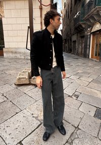 Veste bomber noire avec boutons argentés, t-shirt blanc, jean large en denim foncé et chaussures noires, debout sur une rue pavée.