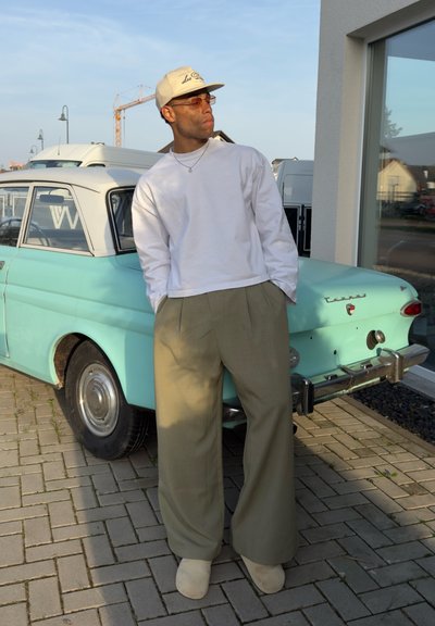 Hombre con gorra beige, camisa blanca de manga larga y pantalones anchos caqui se apoya en un coche antiguo verde menta estacionado en un camino de ladrillos.