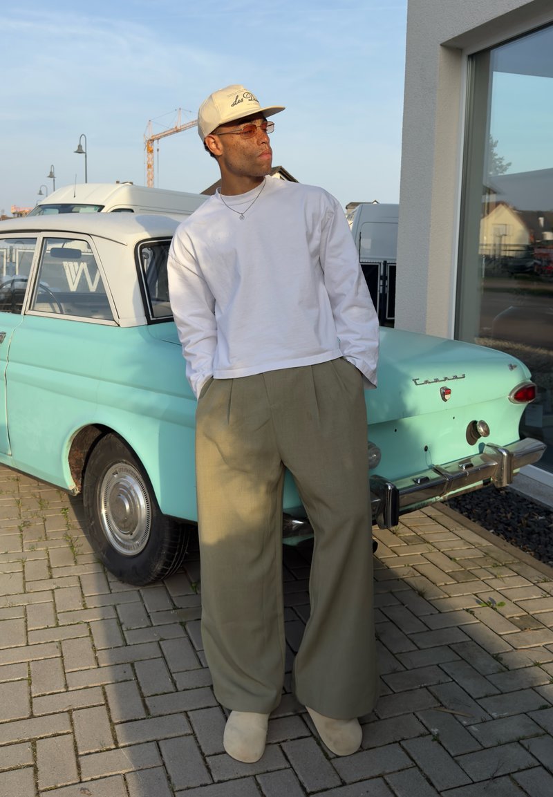 Άνδρας με μπεζ καπέλο, λευκό πουκάμισο μακρυμάνικο και φαρδιά χακί παντελόνια στηρίζεται σε ένα παλαιό αυτοκίνητο χρώματος μέντας που είναι παρκαρισμένο σε λιθόστρωτη είσοδο.