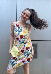 Satin Kleid mit lebhaften floralen und abstrakten Mustern in Rot, Gelb und Blau. Mit verstellbaren Trägern. Hält eine strukturierte gelbe Handtasche.