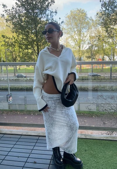 Suéter blanco de gran tamaño con ribete negro, falda de encaje, bolso negro y botas negras de gran tamaño. La modelo está de pie en un balcón con vegetación de fondo.