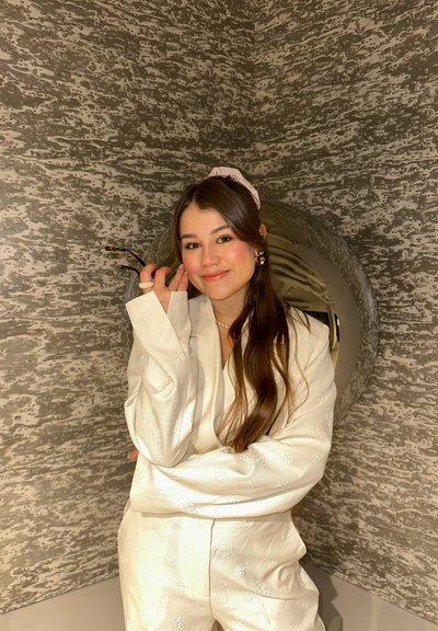 Conjunto de satén blanco con un patrón floral; cabello largo y castaño; lleva un accesorio para el cabello rosa. Fondo con pared gris texturizada.