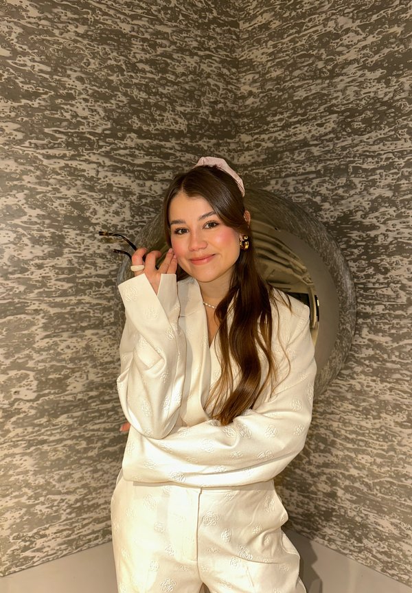 Tenue en satin blanc avec un motif floral ; cheveux longs et bruns ; porte un accessoire pour cheveux rose. Fond avec un mur gris texturé.