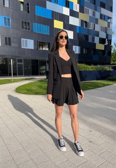 Americana negra sobre un top corto negro ajustado y pantalones cortos negros con pinzas. Combinado con zapatillas negras y blancas; de pie en un camino pavimentado.