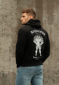 Black hoodie with a front pocket, featuring a large white graphic print of a muscular figure on the back. Worn with blue jeans.
