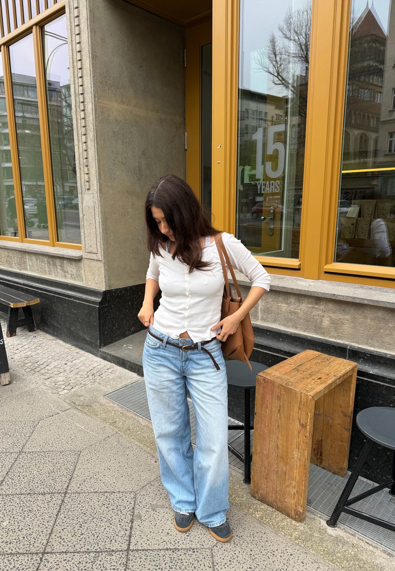 Femme aux longs cheveux foncés portant un haut blanc et un jean bleu ample ajuste sa ceinture devant un bâtiment avec de grandes fenêtres encadrées de jaune et du mobilier en bois.