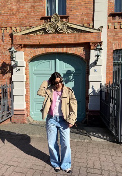 Mujer con gafas de sol, chaqueta beige, suéter rosa y blanco, vaqueros azules, de pie junto a puertas dobles turquesas con ladrillo y ornamentos de piedra.