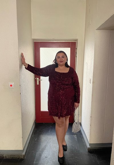 Vestido burdeos con lentejuelas, mangas largas y escote cuadrado, combinado con zapatos de tacón negros y un pequeño bolso plateado, ubicado cerca de una puerta roja.