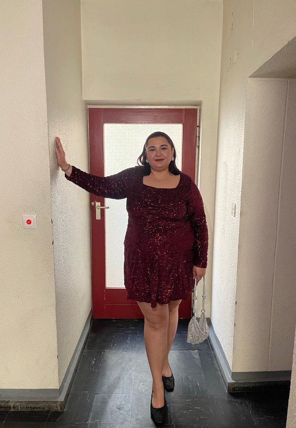 Bordo spalvos suknelė su blizgančiomis detalėmis, ilgomis rankovėmis ir kvadratine iškirpte, derinama su juodais aukštakulniais ir mažute sidabrine rankine, prie raudonų durų.