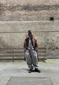 Survêtement gris surdimensionné associé à un manteau en fourrure marron. Accessoire : un bonnet noir. L'arrière-plan présente un mur en béton texturé et un banc en bois.