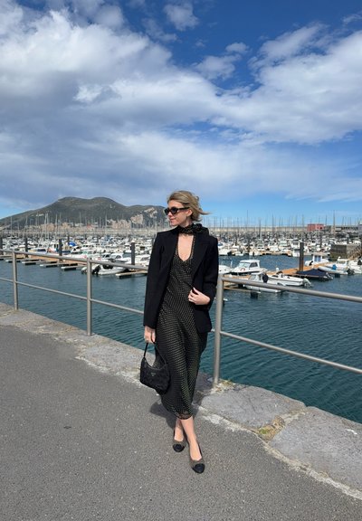 Vestido negro con lunares, escote en V profundo, acompañado de una chaqueta negra y un bolso a juego. El fondo presenta una marina en un día despejado.