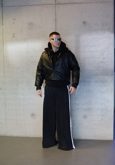 Chaqueta bomber de cuero negra sobre una sudadera con capucha negra, combinada con pantalones anchos negros que tienen rayas laterales blancas. Gafas de sol plateadas y brillantes.