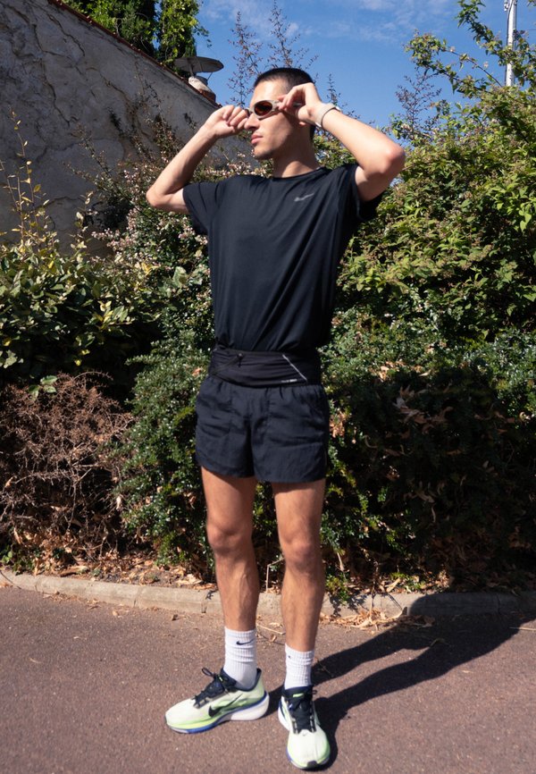 Homme en tenue de sport noire ajustant ses lunettes de soleil tout en se tenant sur un chemin pavé avec des buissons verts et un mur de pierre derrière lui.