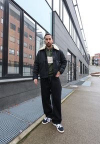 Homme portant une veste noire à col en peau lainée, une chemise zippée verte, un pantalon large noir et des baskets noires et blanches, debout sur un trottoir urbain.