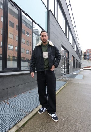 Hombre con chaqueta negra de cuello de borrego, camisa verde con cremallera, pantalones anchos negros y zapatillas negras y blancas, de pie en una acera urbana.