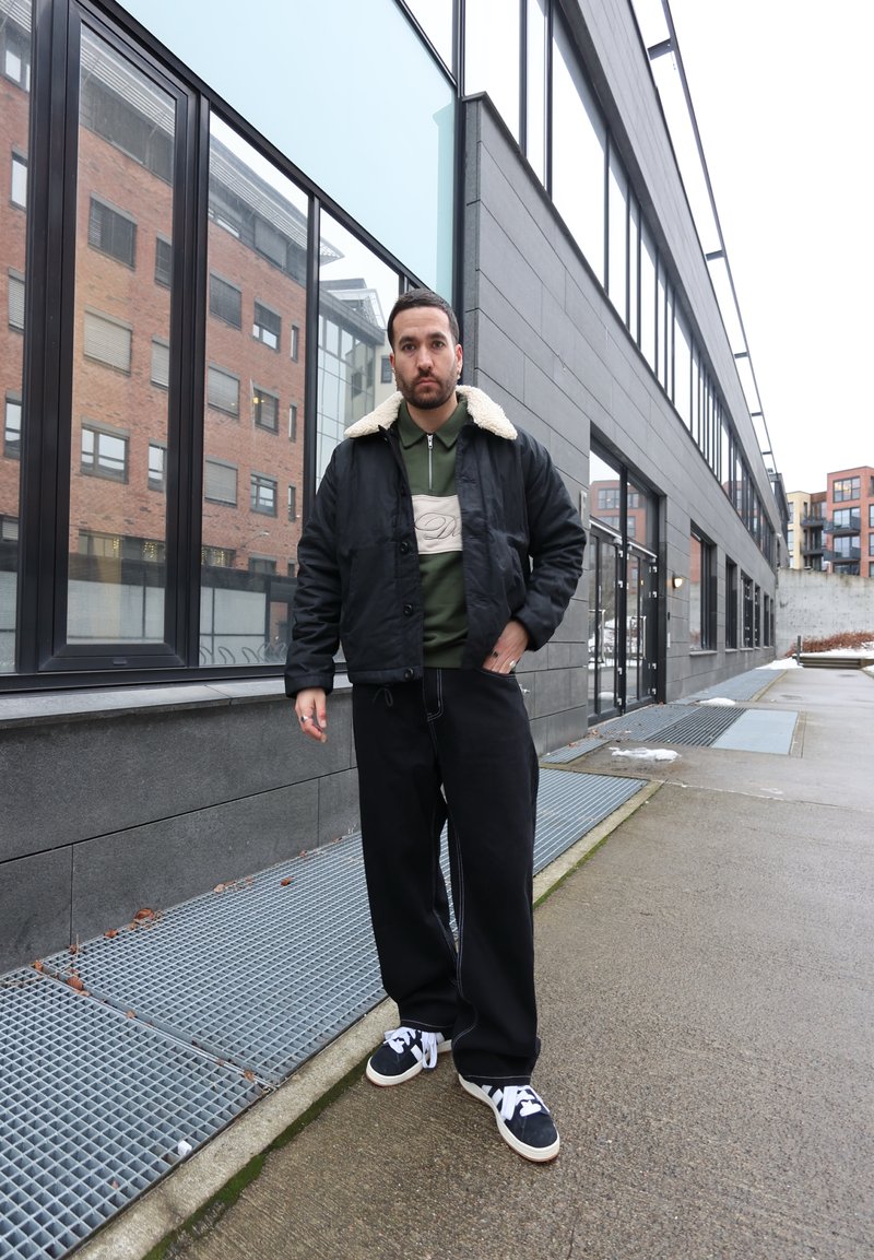 Mann trägt schwarze Jacke mit Lammfellkragen, grünes Reißverschlusshemd, schwarze weite Hose und schwarz-weiße Turnschuhe, steht auf einem städtischen Bürgersteig.