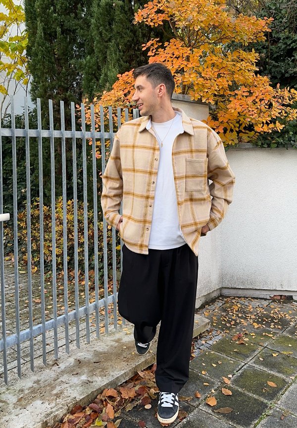 Beige ruudullinen flanellipaita keltaisilla yksityiskohdilla, tyylitelty valkoisen T-paidan päälle. Mustat leveälahkeiset housut ja mustat lenkkarit täydentävät asun.